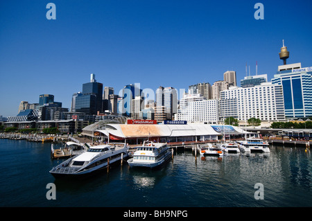 Acquario Darling Harbour Sydney // SYDNEY, Australia - l'acquario di Darling Harbour a Sydney è in primo piano con lo skyline della città sullo sfondo. Il vivace lungomare presenta varie barche attraccate, con la Sydney Tower Eye visibile tra gli edifici torreggianti sotto un cielo azzurro. Foto Stock