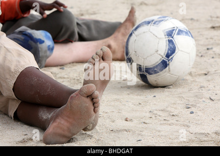 Bambini I piedi sono visibili come essi siedono per terra con un pallone da calcio a guardare un gioco a Khayelitsha, Sud Africa Foto Stock