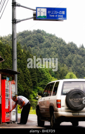 Un uomo acquista un drink da un distributore automatico situato al di sotto di un cartello stradale puntando i visitatori verso la tomba di Cristo in Shingo, Giappone Foto Stock