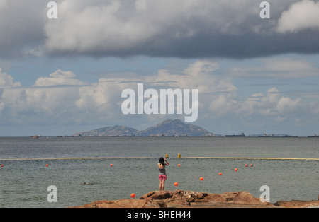 Vista della spiaggia di una donna in piedi sulle rocce, fotografando navi, nel Mare della Cina del Sud, Hung Shing Yeh, Lamma, Isola di Hong Kong Foto Stock
