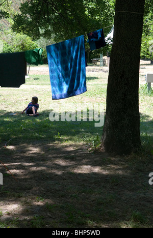 7 anno vecchio ragazzo giovane con frisbee siede su erba in ombra al di sotto di una linea di lavaggio con asciugamani di essiccazione in un campeggio in Francia Foto Stock