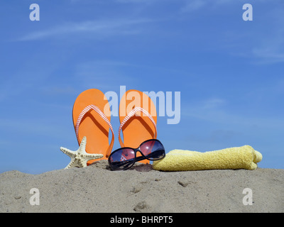 Orange flip-flop azionati verticalmente in spiaggia di sabbia, stelle marine, occhiali da sole e giallo asciugamano da bagno oltre il cielo blu Foto Stock