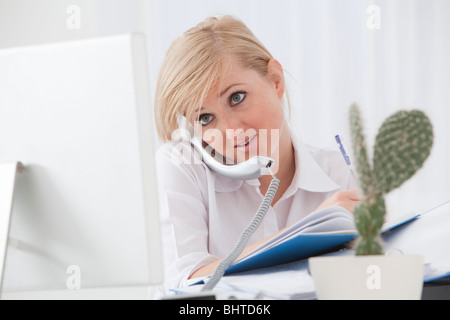 Occupato il multitasking giovane donna di fronte allo schermo del computer e del telefono alla sua scrivania in un moderno ufficio Foto Stock