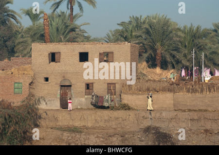 Vista di case sulla sponda del fiume Nilo, nei pressi di Luxor in Egitto con la popolazione locale in abiti colorati e una donna che trasportano l'acqua Foto Stock