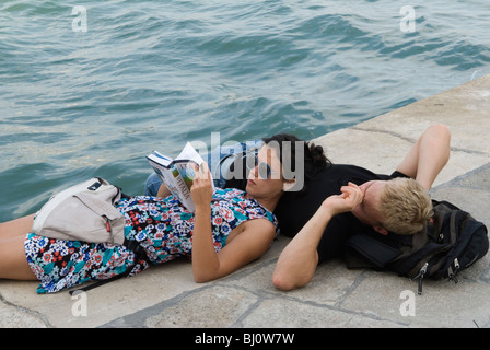 Giovani coppie adulte innamorate che si divertono a vicenda in vacanza a Venezia. Sdraiati sul lato di un canale esausti, stanchi... Sta leggendo un libro di guida di viaggio in italiano. HOMER SYKES anni '2009 2000 Foto Stock