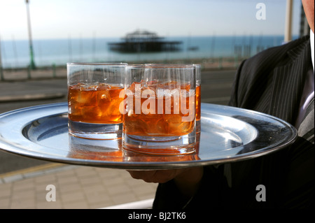 Tumbler bicchieri di whisky con grumi di cubetti di ghiaccio su un vassoio tenuto a mano nella parte anteriore del Molo Ovest di Brighton Seafront Foto Stock