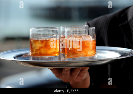 Tumbler bicchieri di whisky con grumi di cubetti di ghiaccio su un vassoio tenuto a mano nella parte anteriore del Molo Ovest di Brighton Seafront Foto Stock