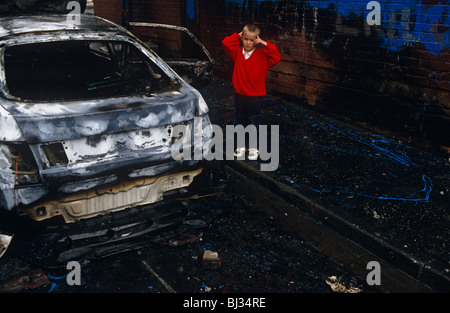 Un giovane ragazzo guarda traumatizzati in piedi accanto a un bruciate le shell di un automobile berlina incendiata dai vandali a Belfast. Foto Stock