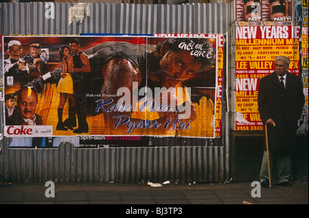 Un uomo anziano di discesa South-Asian sta aspettando un autobus a Southall vicino a Bollywood manifesti di cinema. Foto Stock