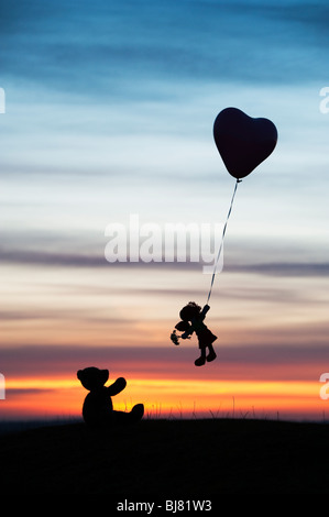 Rag Doll tenuta su di una forma di cuore palloncino elio che si allontanano da un orsacchiotto di sunrise. Regno Unito. Ancora vita silhouette Foto Stock