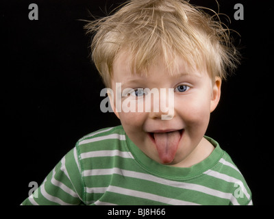 Little Boy che mostra la sua lingua su sfondo nero. Ragazzo hanno gli occhi blu, capelli biondi e un po' di sporcizia sul viso Foto Stock