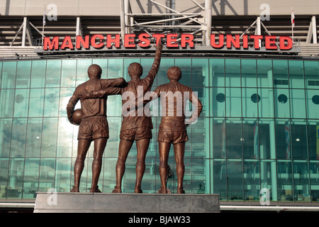 La statua di Sir Bobby Charlton, George Best Denis legge presso l'ingresso principale (Supporto EST) a Old Trafford, Manchester. Foto Stock