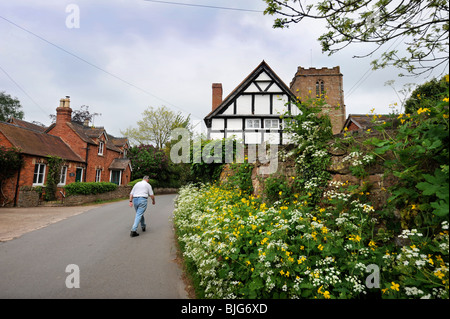 Un uomo che cammina verso il basso di una corsia di marcia nel villaggio di Redmarley D'Abitot, Gloucestershire con la Chiesa di San Bartolomeo del Foto Stock