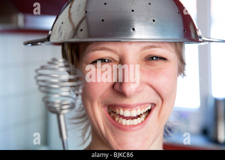 Donna sorridente in una cucina con un setaccio da cucina su di lei e una frusta in mano la riproduzione di un duello di cottura Foto Stock