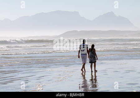 Giovane camminando mano nella mano sul bagnato spiaggia sabbiosa a trefolo nel Western Cape Sud Africa nella distanza Table Mountain Foto Stock