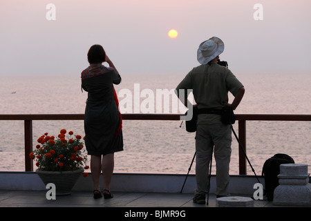 Donna di prendere una fotografia e un fotografo con una telecamera su un treppiede di fronte al tramonto sul mare, Kovalam, Kerala, sout Foto Stock