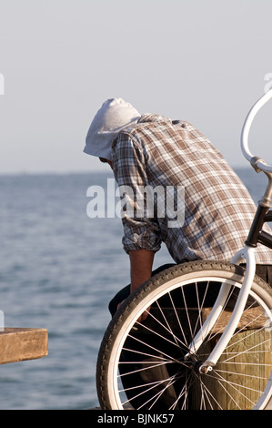 Giovane uomo con la felpa con cappuccio e plaid shirt seduta sul banco di molo vicino oceano Foto Stock