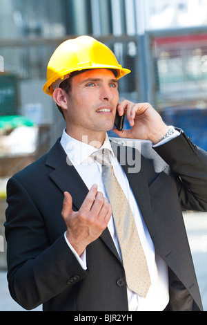 Un uomo in un casco giallo e tuta su scala industriale o sito in costruzione a parlare sul suo telefono cellulare Foto Stock