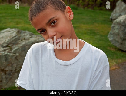 Questo closeup ritratto è un grazioso 8 anno vecchio bi-razziale ragazzo indossa plain white T-shirt e fuori con erba e alcune rocce di grandi dimensioni Foto Stock