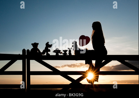 Ragazza con un cuore palloncino con una rag doll, pollo, la volpe, il coniglio e orso peluche seduto su un cancello al tramonto. Silhouette Foto Stock