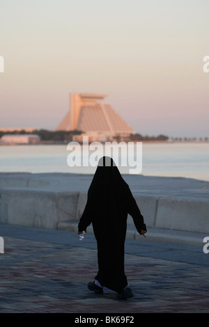 La donna araba che indossano il tradizionale abito nero a piedi lungo la Corniche, Sheraton Hotel nel retro, a Doha, in Qatar Foto Stock