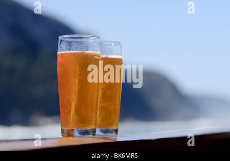 Due bicchieri di birra su una ringhiera, con una spettacolare costa come sfondo. Focus sui bicchieri Foto Stock