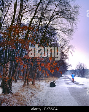 Il jogger solitario del parco innevato si fa strada lungo un sentiero invernale ghiacciato e innevato nel lago Virginia Water, Surrey, Inghilterra, Regno Unito Foto Stock