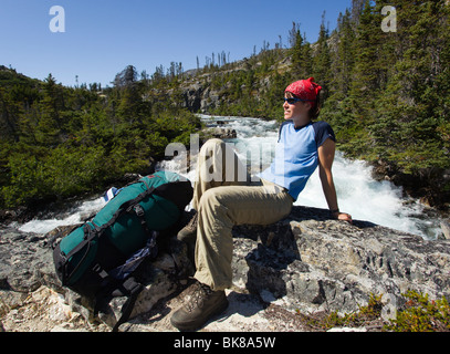 Giovane donna, escursionista backpacker, zaino, seduto sulla roccia, riposo, Moose Creek Canyon, cascata dietro, storico Chilkoot Pas Foto Stock