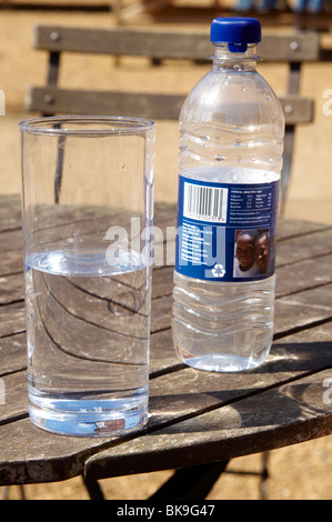 Bicchiere di acqua con bottiglia d'acqua in plastica dietro in piedi su un a doghe di legno e rabboccato tavolo da picnic. Foto Stock