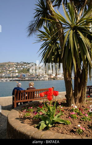 Coppia di anziani seduti su un sedile di legno sulla banchina a Dartmouth & affacciato Kingswear e fiume Dart South Devon England Foto Stock