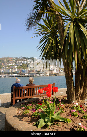 Coppia di anziani seduti su un sedile di legno sulla banchina a Dartmouth & affacciato Kingswear e fiume Dart South Devon England Foto Stock