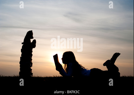 Ragazza giovane disteso la lettura di un libro di fronte a un orsacchiotto seduto su una pila di libri al tramonto. Silhouette Foto Stock