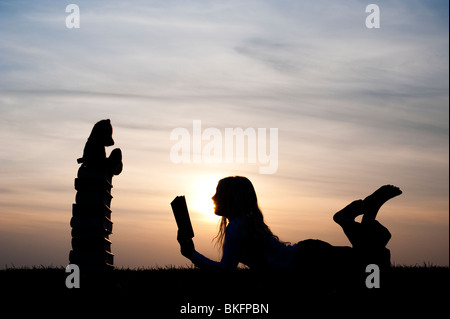 Ragazza giovane disteso la lettura di un libro di fronte a un orsacchiotto seduto su una pila di libri al tramonto. Silhouette Foto Stock