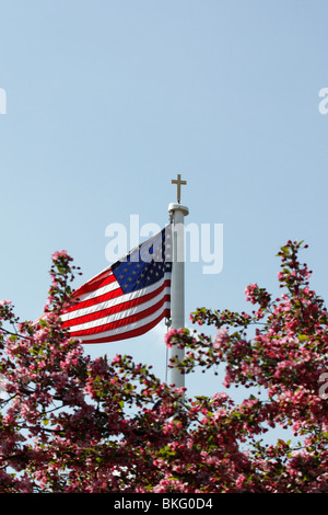 La chiesa cattolica con una croce e una bandiera americana su un palo con albero fiorito contro il cielo blu angolo basso nessuno verticale in Ohio Stati Uniti alta risoluzione Foto Stock