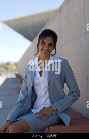 Una giovane e bella (orientale) donna indiana che si siede su una panchina lungo una parete di cemento che indossa casual abbigliamento aziendale e sorridente. Foto Stock