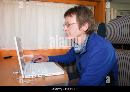 Donna matura del baby boom generazione utilizzando il wifi su un laptop MacBook computer all'interno di un camper mentre camping. Inghilterra, Regno Unito, Gran Bretagna Foto Stock