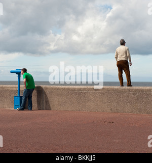 Vista posteriore di un uomo in piedi sulla parete del mare, un altro uomo guardando attraverso il telescopio, Rhyl, north Wales UK Foto Stock