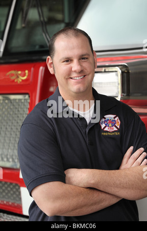 Bello vigile del fuoco nel suo ventenne con le braccia incrociate in piedi di fronte a firetruck close-up Foto Stock