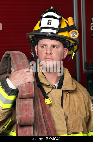 Bel giovane vigile del fuoco tenendo tubo antincendio in uniforme di fronte firetruck Foto Stock