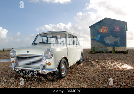 Un bianco Austin Mini Cooper mk2 la macchina parcheggiata su una spiaggia di ciottoli sulla costa sud con un pesce-verniciato beach hut in background. Foto Stock