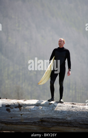 Un uomo si erge su un albero caduto con la sua tavola da surf sulla costa perso, California. Foto Stock
