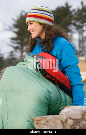 Una giovane donna entra nel suo sacco a pelo come si siede sul driftwood in una foresta aperta impostazione. Foto Stock