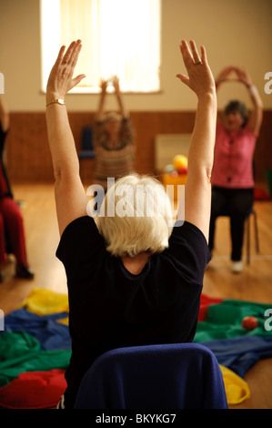 Vista posteriore del gruppo dei cittadini anziani che partecipano a un basso impatto sedia lezione di aerobica in un piccolo villaggio hall in West Wales UK Foto Stock