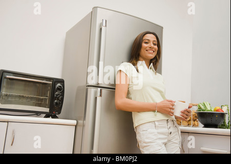 Donna appoggiata contro un frigorifero e tenendo una tazza da caffè Foto Stock