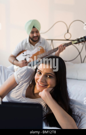 Donna che utilizza un computer portatile e di suo marito a suonare la chitarra sul letto Foto Stock