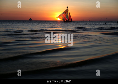 Una imbarcazione a vela passa il tramonto sulla spiaggia bianca, Boracay, la più famosa destinazione turistica nelle Filippine. Foto Stock