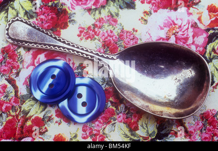 Chiusura del vecchio silver tea caddy cucchiaio con due pulsanti blu su rosa materiale floreale sfondo Foto Stock