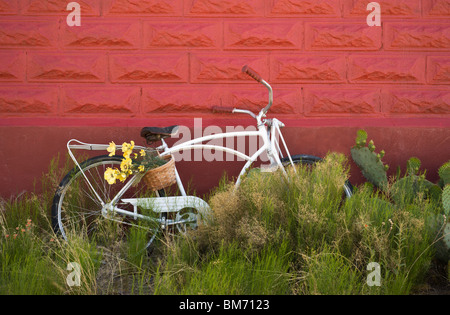 Una vecchia bicicletta abbandonata, parcheggiato in erbacce, si appoggia contro la parete del mattone rosso in tre fiumi, Nuovo Messico. Foto Stock