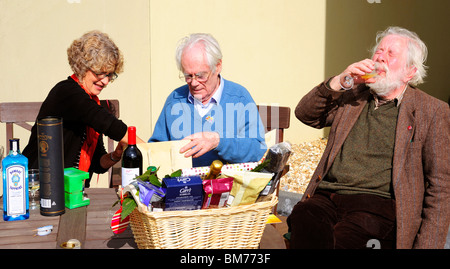 Una giovane donna aiuta un vecchio uomo aperto presenta in occasione del suo novantesimo compleanno mentre un altro vecchio uomo beve vino. Foto Stock