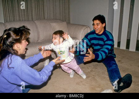 Baby giggles e risate come lei prende i primi passi tra la madre e il padre, mamma e papà figlia orgogliosa divertente Foto Stock